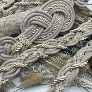 Könnte beinhalten: Beige und goldfarbenes geflochtenes Seilband mit einem dekorativen floralen Design. Das Band ist mit einem dicken Seil geflochten und hat einen zarten goldenen Faden.