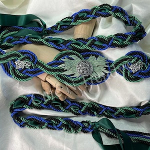 May include: A decorative braided belt in green, black, and blue. The belt features a central medallion and silver-toned embellishments. The design incorporates satin ribbon and rope-like braiding.