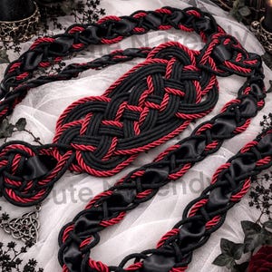 May include: A decorative belt crafted from black and red braided rope intertwined with black satin ribbon. The center features an intricate woven knot design, and a silver-toned metal triquetra charm with a small red stone hangs from one end.