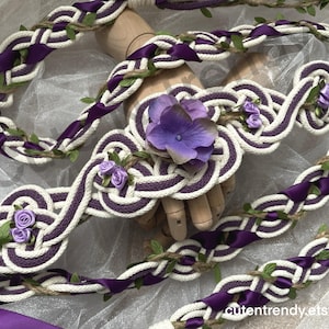 May include: A braided belt featuring white rope intertwined with deep purple satin ribbon and small green leaf accents. The center displays a large purple hydrangea flower and smaller purple rose buds. The belt is displayed on a wooden hand form.