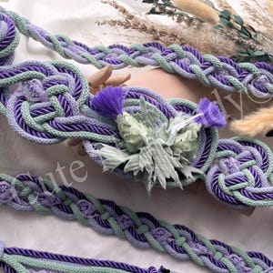 May include: A decorative braided belt in purple and green, with detailed knotwork and a floral embellishment. The belt is crafted from rope and is suitable for various outfits. The design includes a thistle-like flower, adding a touch of nature-inspired elegance.