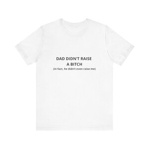 Puede incluir: Una camiseta blanca de manga corta con texto negro impreso en el pecho. El texto dice: DAD DIDN'T RAISE A BITCH (in fact, he didn't even raise me). La prenda tiene un cuello redondo y un corte estándar, confeccionada en tela blanca lisa.