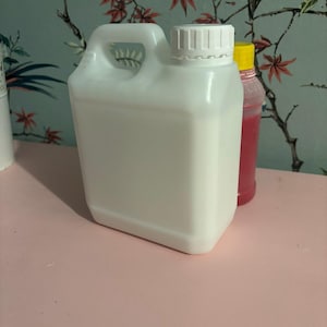May include: A white plastic jug with a handle and a screw-on cap. The jug is rectangular with rounded corners. A red and yellow bottle is visible behind the jug. The background features a floral wallpaper.