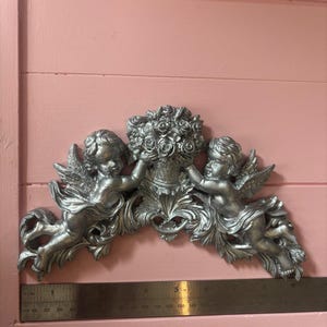 May include: Silver-colored cherub wall decor featuring two winged figures holding a floral arrangement. The decorative piece is approximately 6 inches wide, with intricate details and a classical design.