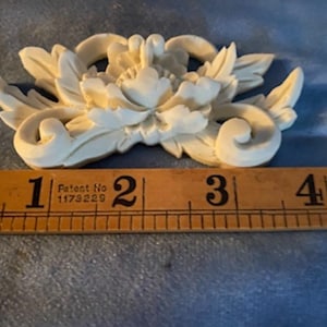May include: Off-white floral decorative element with a detailed design of leaves and petals. The piece is approximately 4 inches long, as measured by a wooden ruler. The item has a vintage aesthetic and is set against a blue background.