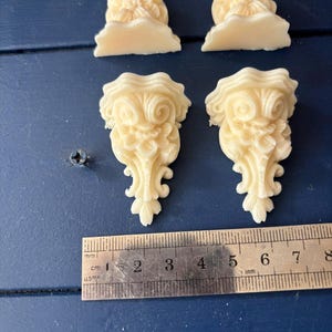 Op de afbeelding: Vier crèmekleurige decoratieve architecturale ornamenten. Twee zijn bloemvormig en twee zijn rolvormig. Een liniaal toont de afmetingen in centimeters. De ornamenten liggen op een donkerblauwe ondergrond.