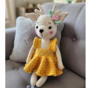 Könnte beinhalten: Handgefertigte Häkeltierpuppe, cremefarbener Körper, gelbes Kleid. Das Reh hat eine schwarze Nase, rosa Wangen und einen Blumenschmuck. Die Puppe sitzt auf einem grauen Sofa.