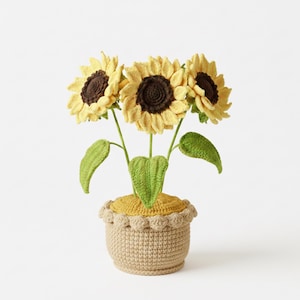Könnte beinhalten: Ein handgefertigtes gehäkeltes Sonnenblumenarrangement in einem beigen Topf. Die Sonnenblumen haben gelbe Blütenblätter und braune Zentren. Grüne Blätter und Stiele tragen die Blumen. Der Topf hat einen gewellten Rand und einen gelben Boden.
