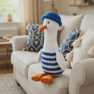 Könnte beinhalten: Eine handgefertigte Häkelgans-Puppe mit blauem Matrosenhut und blau-weiß gestreiftem Hemd. Die Gans hat einen orangefarbenen Schnabel und Füße. Die Puppe sitzt auf einem weißen Sofa mit blau gemusterten Kissen.