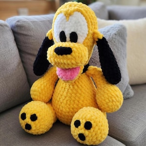 May include: A handmade yellow plush dog with black ears, nose, and spots, and a pink tongue. The stuffed animal is sitting on a grey sofa. The dog's eyes are white. The plush toy is a cute, cuddly item.