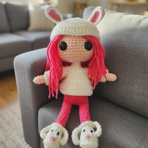 Könnte beinhalten: Handgemachte Häkelpuppe mit weißem Hasenhut mit rosa Ohren, rosa Haaren, einem weißen Pullover und einer rosa Hose. Die Puppe hat schwarze Augen und trägt weiße Hasenhausschuhe mit rosa Nasen.