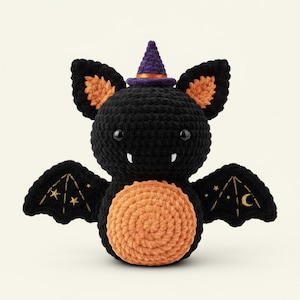 Könnte beinhalten: Eine handgefertigte schwarze Häkel-Fledermaus mit orangefarbenen Akzenten, die einen lila Hexenhut trägt. Die Fledermaus hat bestickte Flügel mit goldenen Sternen und einem Mond. Der Bauch der Fledermaus ist orange und hat kleine weiße Zähne.