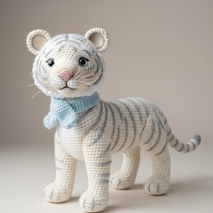 Könnte beinhalten: Handgefertigtes gehäkeltes Tiger-Spielzeug in Creme und Grau, mit einem hellblauen Schal. Der Tiger hat graue Streifen, eine rosa Nase und schwarze Augen. Die Ohren sind cremefarben. Ein bezauberndes, weiches Spielzeug.