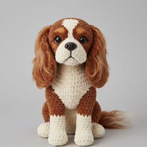 Könnte beinhalten: Ein gehäkelter Cavalier King Charles Spaniel Hund mit braunen, weißen und hellbraunen Farben. Der Hund hat lange, schlaffe Ohren, eine weiße Brust und weiße Pfoten. Der Hund sitzt aufrecht und hat ein weiches, strukturiertes Aussehen.