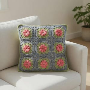 Könnte beinhalten: Quadratisches Häkelkissen mit Blumenmuster. Das Kissen hat einen grauen Hintergrund mit neun quadratischen Blumendesigns in Rosa und Grün. Das Kissen hat einen grünen Rand und liegt auf einem weißen Sofa.