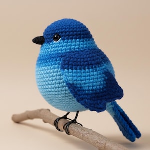 Könnte beinhalten: Ein handgehäkelter Vogel in Blautönen, der auf einem kleinen Ast sitzt. Der Vogel hat einen schwarzen Schnabel und Augen, wobei verschiedene blaue Garne ein strukturiertes Aussehen erzeugen. Der Vogel ist etwa 15 cm lang.