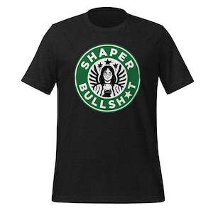 May include: Dark gray t-shirt with a green and white circular graphic. The graphic features a woman with glasses and the words "SHAPER BULLSH*T" in a Starbucks-inspired design. The shirt is short-sleeved.