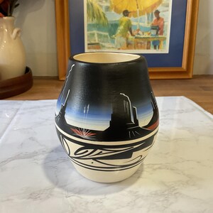 May include: A black and cream-colored ceramic vase with a landscape design. The vase features a black upper portion with a painted desert scene, including red, white, and blue accents. The bottom half is cream-colored with a decorative black and white pattern.