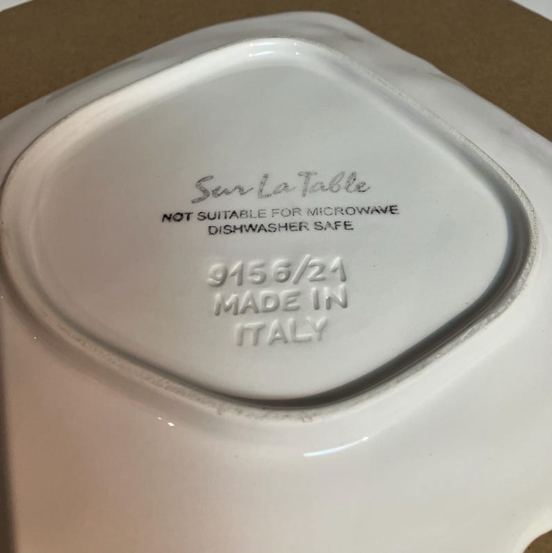 Peut inclure: Le dessous d'un plat en c&eacute;ramique blanc portant l'inscription Sur La Table, NOT SUITABLE FOR MICROWAVE, DISHWASHER SAFE, 9156/21 et MADE IN ITALY grav&eacute;e et imprim&eacute;e &agrave; l'encre noire au centre de la base de forme carr&eacute;e.