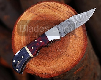 May include: A Damascus steel folding knife with a patterned blade and a handle featuring red, black, and gold accents. The knife is resting on a cross-section of a tree trunk. The text "Blades Barn" is visible.