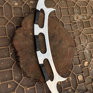 Könnte beinhalten: Eine silberfarbene, gebogene Klinge mit drei spitzen Kanten und einem schwarzen Griff. Die Klinge hat ein einzigartiges, symmetrisches Design und wird auf einer Holzoberfläche präsentiert.