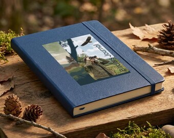 Puede incluir: Un diario azul con el texto "Birders Journal" y una imagen de un observador de aves y un águila. El diario está sobre una superficie de madera con musgo, piñas y pequeñas piedras.