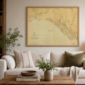 May include: A large rectangular wall art print featuring a vintage-style map of Alaska with a yellow-toned background, black cartographic details, and text reading Alaska, displayed in a simple light-coloured wooden frame on a plain wall.