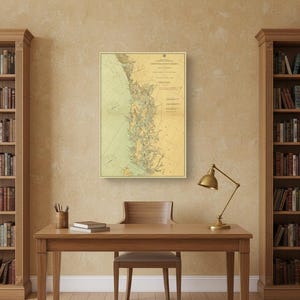 May include: A framed vintage nautical chart, with a detailed map in green and yellow tones. The chart is mounted on a wall between two bookshelves. A desk with a lamp and chair is in front of the chart.