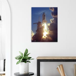 May include: A canvas print of a space shuttle launching into a blue sky filled with clouds. The shuttle is white with black accents, ascending with a bright orange flame. The launch tower is visible in the foreground.