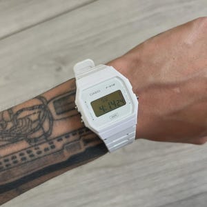 May include: A white Casio F-91W digital watch is shown on a wrist. The watch face displays the time as 4:14:24. The watch has a white strap and a rectangular face with black digital numbers. The watch is water resistant.