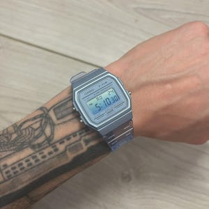 Puede incluir: Un reloj digital plateado Casio F-91W que muestra la hora 5:10:30. El reloj tiene una esfera rectangular con una pantalla digital y una correa plateada. El reloj está en una muñeca.