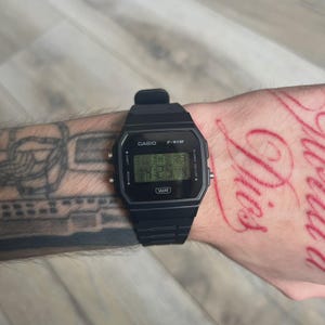May include: A black Casio F-91W digital watch with a rectangular face and a black strap. The watch shows the time on a green digital display. The watch has the Casio logo and the letters "WR" on the face.