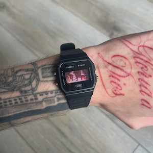 May include: Black Casio F-91W digital watch showing the time 14:37:12. The watch has a rectangular face and a black resin strap. The watch is on a wrist with tattoos.