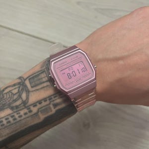 Pode incluir: Um relógio digital rosa Casio F-91W exibindo a hora 8:01:10. O relógio tem um mostrador retangular e uma pulseira rosa combinando. O relógio está em um pulso.
