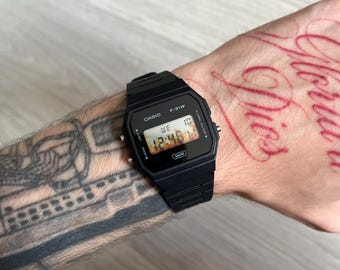 May include: A black digital wristwatch with a rectangular face and a matching black resin strap. The watch display shows the text CASIO and F-91W at the top, with the time 12:46 and the day WE 10 in the center, and the letters WR at the bottom.
