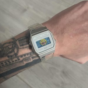 Peut inclure: Une montre numérique Casio F-91W argentée avec un bracelet en plastique transparent. Le cadran affiche l'heure et un graphique. La montre est portée au poignet.