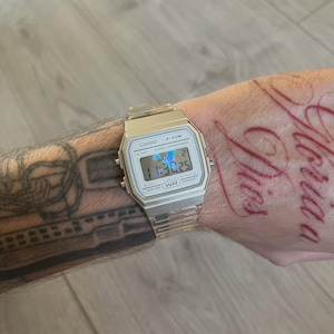 May include: Silver Casio F-91W digital watch with a clear strap. The watch face shows the time, date, and day. The watch is water-resistant. The watch is on a wrist with tattoos.