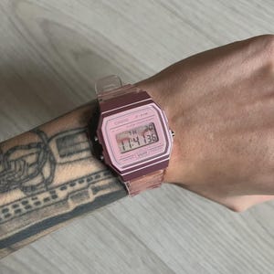 May include: A pink Casio F-91W digital watch with a clear band is displayed on a wrist. The watch face shows the time, date, and day. The watch has a rectangular face and a clear plastic band.