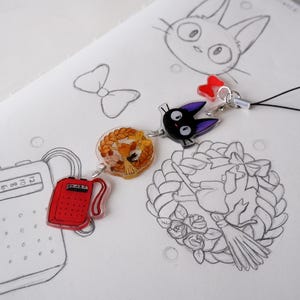 May include: A selection of anime-themed charms and sketches. The charms include a black cat with purple ears, a charm with a character inside a wreath, and a red radio. The sketches feature a cat, a bow, and a witch on a broom.