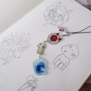 May include: A detailed charm with anime-inspired designs. The charm features a red and white circular pendant, a small white figure, and a blue and white pendant. The charm is attached to a black cord and rests on a sketchbook with pencil sketches.
