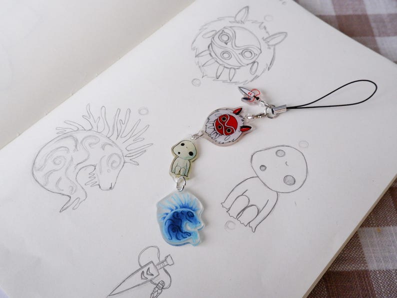 May include: A detailed charm with anime-inspired designs. The charm features a red and white circular pendant, a small white figure, and a blue and white pendant. The charm is attached to a black cord and rests on a sketchbook with pencil sketches.