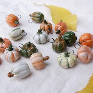May include: A collection of small, glazed ceramic pumpkin charms in various colors including orange, white, and dark green. Each miniature pumpkin features a small metal loop at the top for hanging, with a glossy finish and distinct ridges.