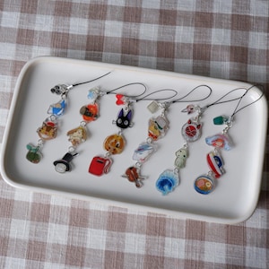 Puede incluir: Una colección de coloridos amuletos de estilo anime con cordones negros, exhibidos en una bandeja rectangular blanca. Los amuletos presentan varios personajes y objetos de dibujos animados, incluyendo un gato, un sombrero y una bolsa roja.