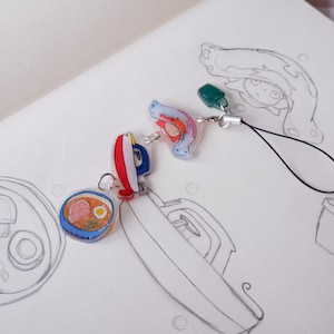 May include: A selection of colourful anime-style charms, including a ramen bowl, a red and blue boat, and a character with red hair. The charms are attached to a black lanyard and are set against a white background with pencil sketches.