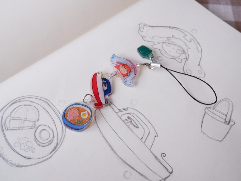 May include: A selection of colourful anime-style charms, including a ramen bowl, a red and blue boat, and a character with red hair. The charms are attached to a black lanyard and are set against a white background with pencil sketches.