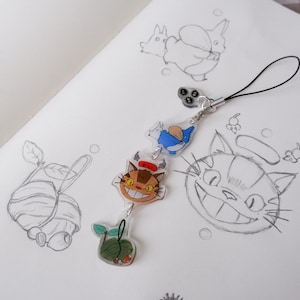 May include: A clear acrylic charm featuring three colourful Studio Ghibli characters: a cat, a blue creature, and a green plant. The charm is attached to a black lanyard, set against pencil sketches.