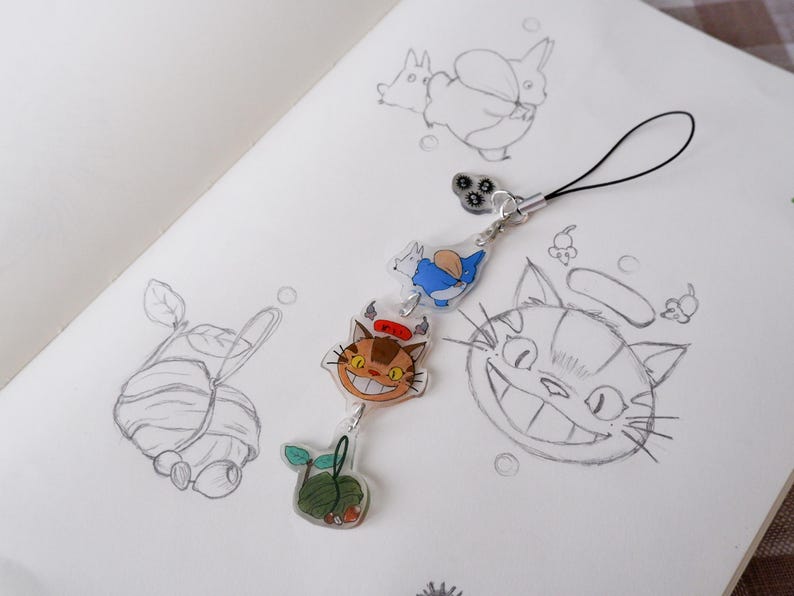 May include: A clear acrylic charm featuring three colourful Studio Ghibli characters: a cat, a blue creature, and a green plant. The charm is attached to a black lanyard, set against pencil sketches.