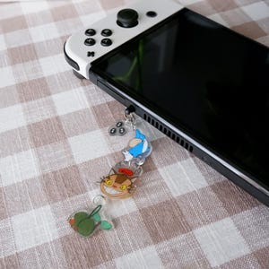 May include: A black and white Nintendo Switch console with a colourful charm featuring cartoon characters. The charm includes a blue creature, a smiling cat, and a green leaf. The console rests on a checkered surface.