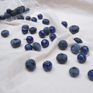 Puede incluir: Una colección de pequeñas cuentas redondas de color azul oscuro con forma de arándano, cada una con una pequeña anilla metálica en la parte superior. Algunas de estas piezas están esparcidas sobre una tela blanca, mientras que otras permanecen dentro de un recipiente de plástico transparente.