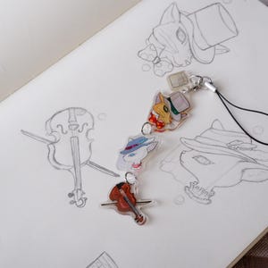 May include: A selection of cat-themed charms and a miniature violin pin are displayed on a sketchbook page with pencil sketches. The charms depict cats wearing hats and bow ties, with a clear, glossy finish. The violin pin is reddish-brown.